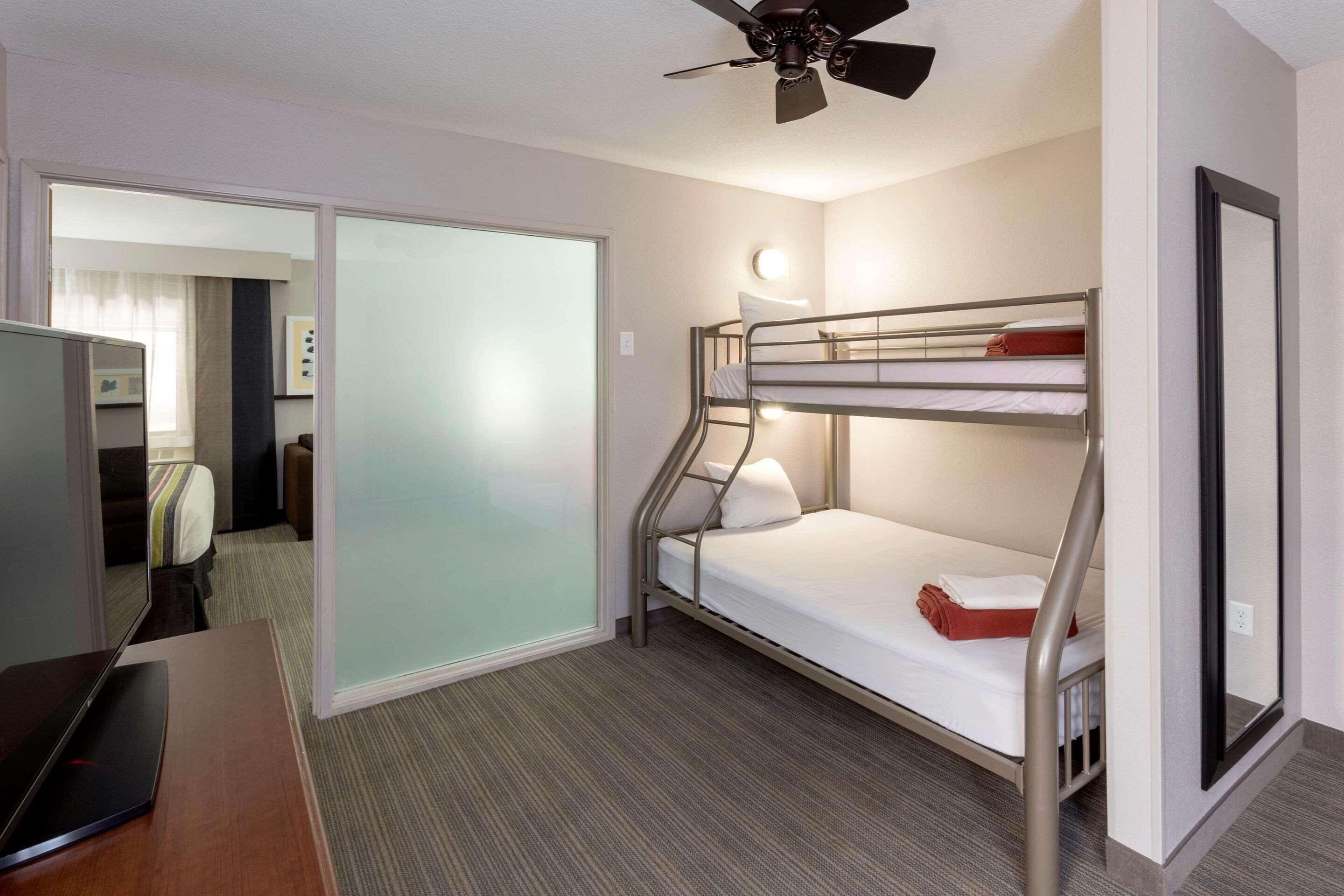 Une chambre d’hôtel avec un lit superposé, un bureau avec un ordinateur portable, un ventilateur de plafond et une porte coulissante donnant sur une autre chambre.