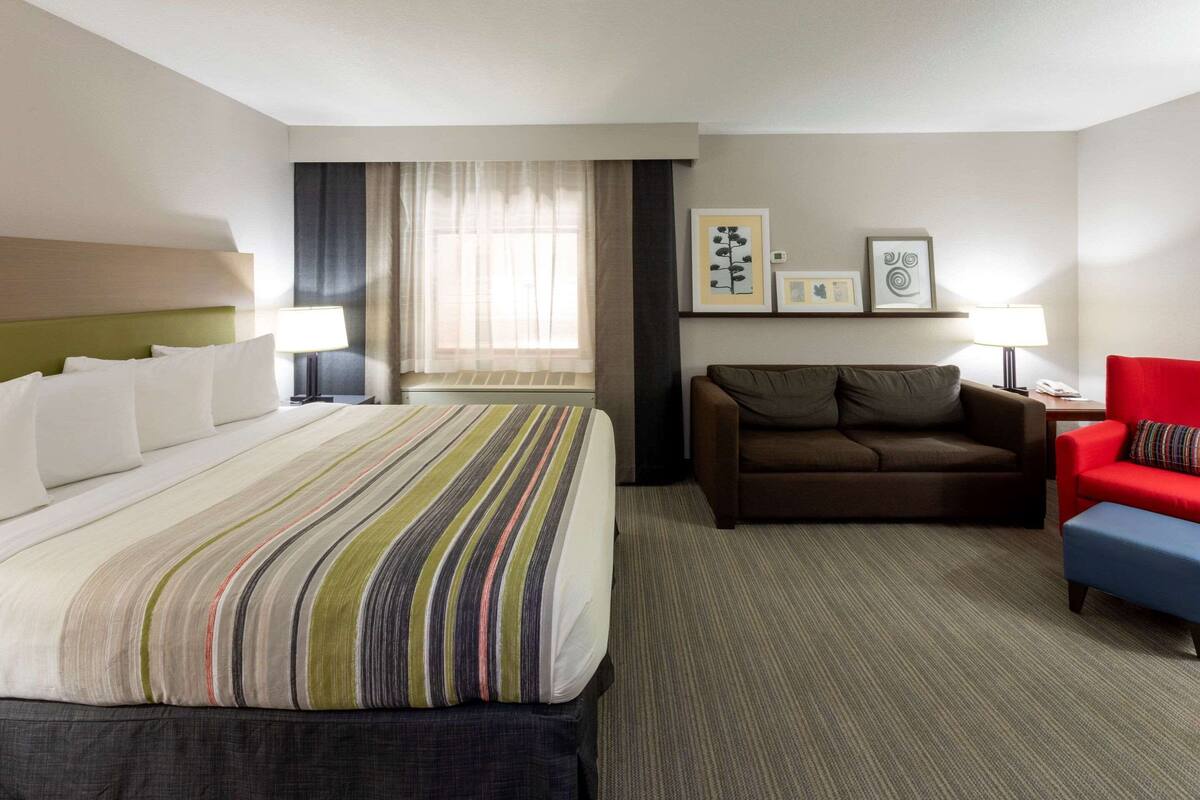 A hotel room with a bed, a sofa, a red armchair, a nightstand, and framed pictures on the wall.