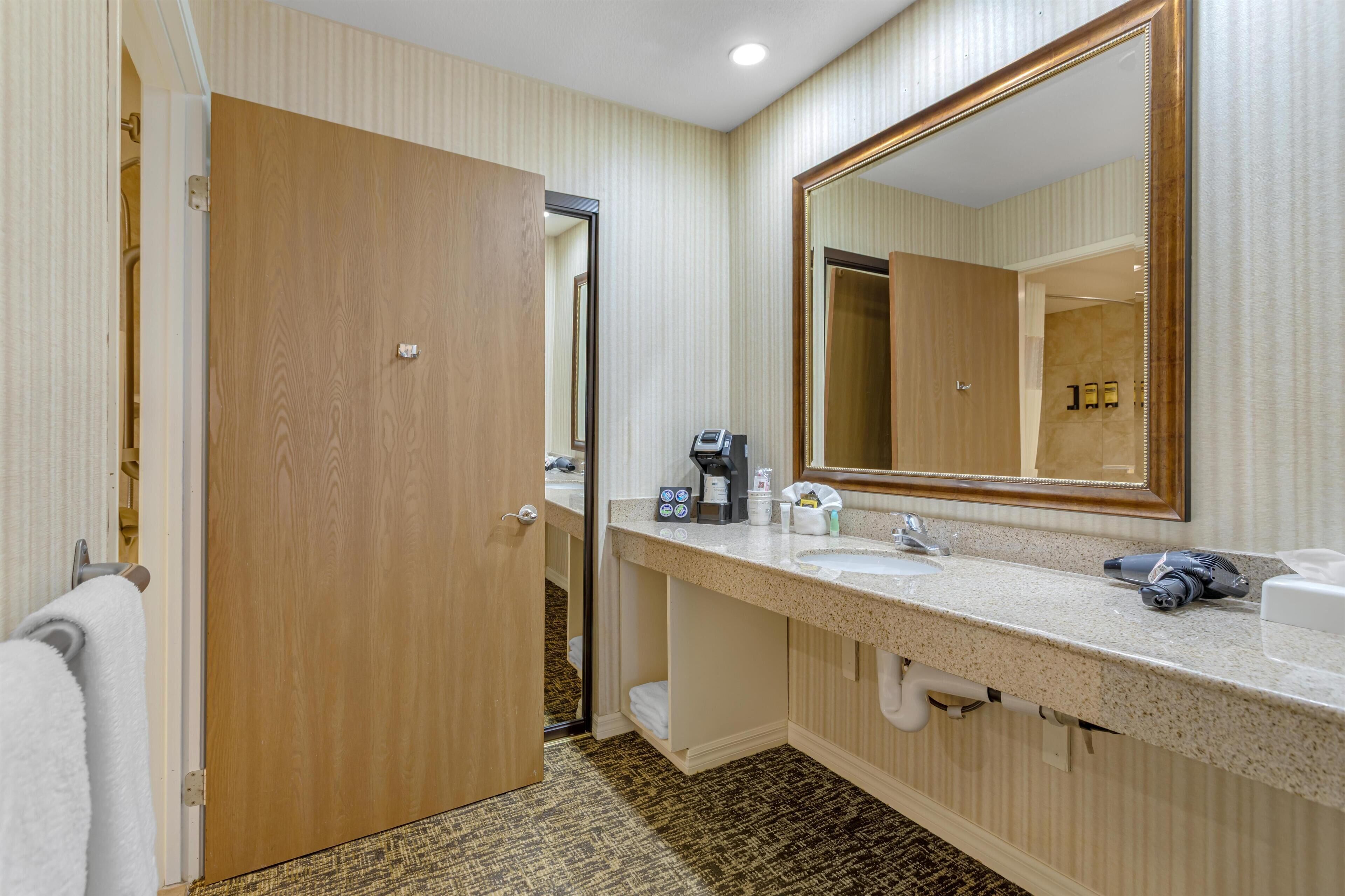 A hotel bathroom with a wooden door, a large mirror, a countertop with a coffee maker, and a sink.