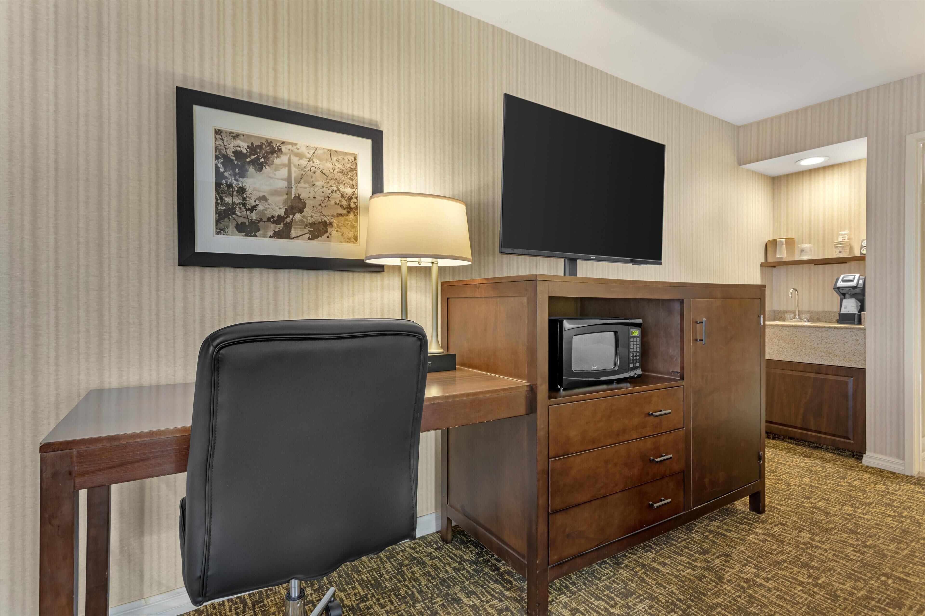 Ein Hotelzimmer mit Schreibtisch, Stuhl, Fernseher, Mikrowelle und einer kleinen Küchenzeile.