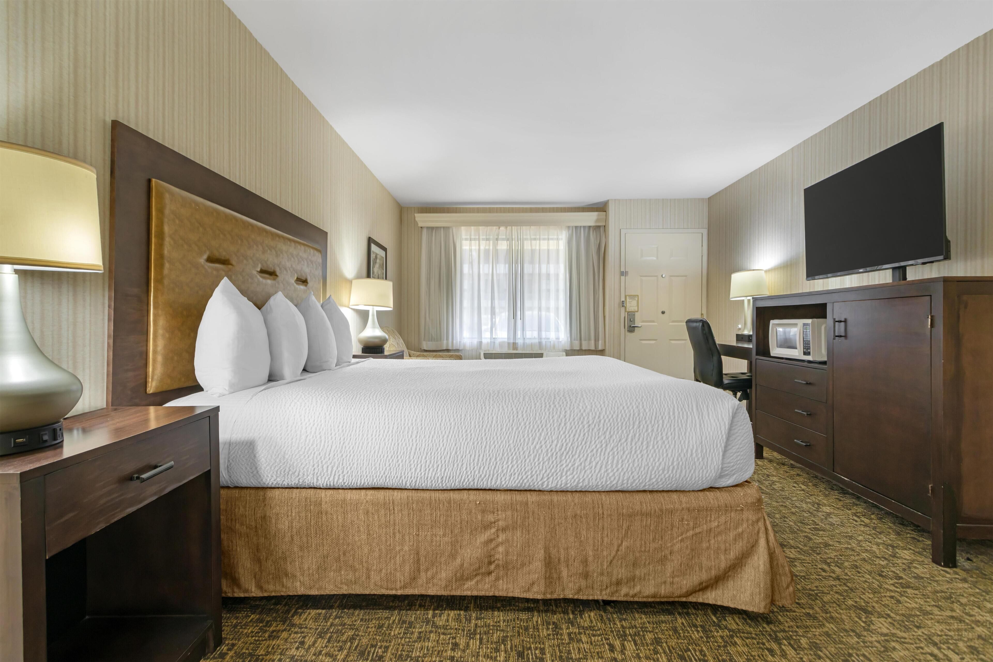 A hotel room with a large bed, a flat-screen TV, a desk with a lamp, a wardrobe, and a window with curtains.
