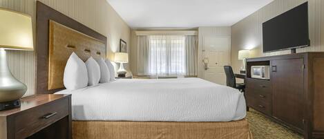A hotel room with a large bed, a flat-screen TV, a desk with a lamp, a wardrobe, and a window with curtains.