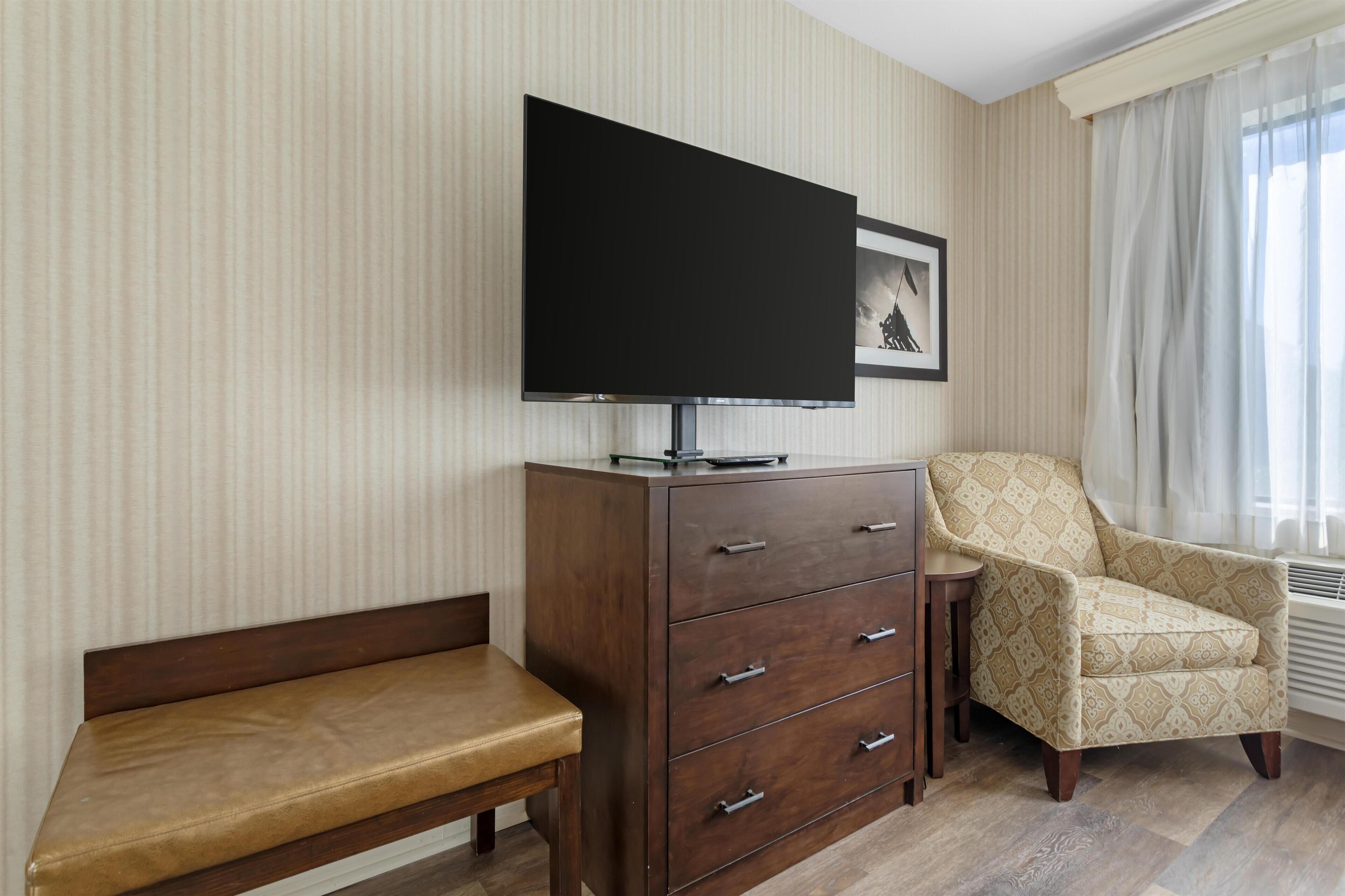 Una habitación con una televisión de pantalla plana sobre un mueble de madera, un sillón estampado, un banco y un cuadro en la pared.