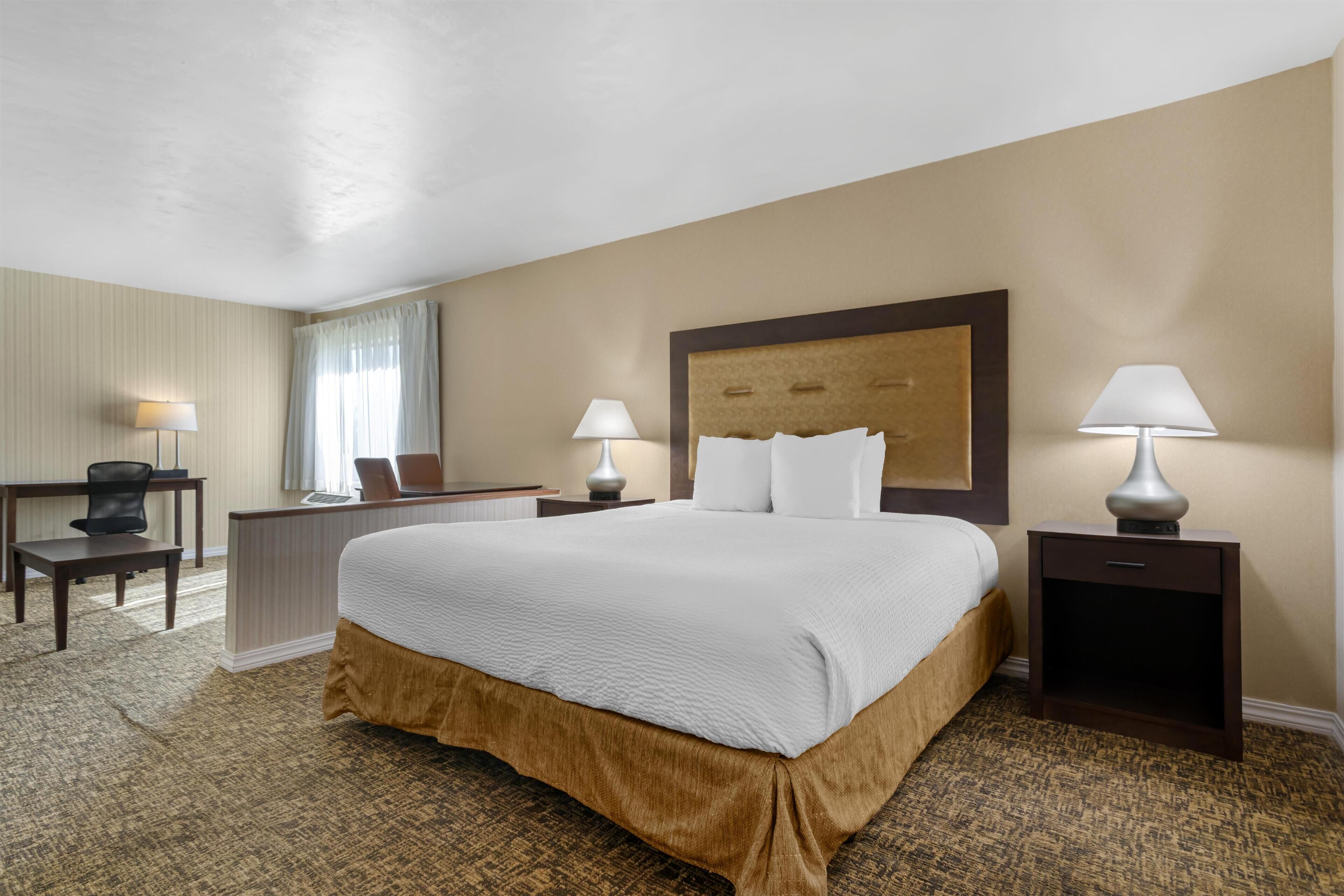 Quarto de hotel com uma cama grande, duas mesas de cabeceira com luminárias, uma escrivaninha com cadeira e uma janela com cortinas.