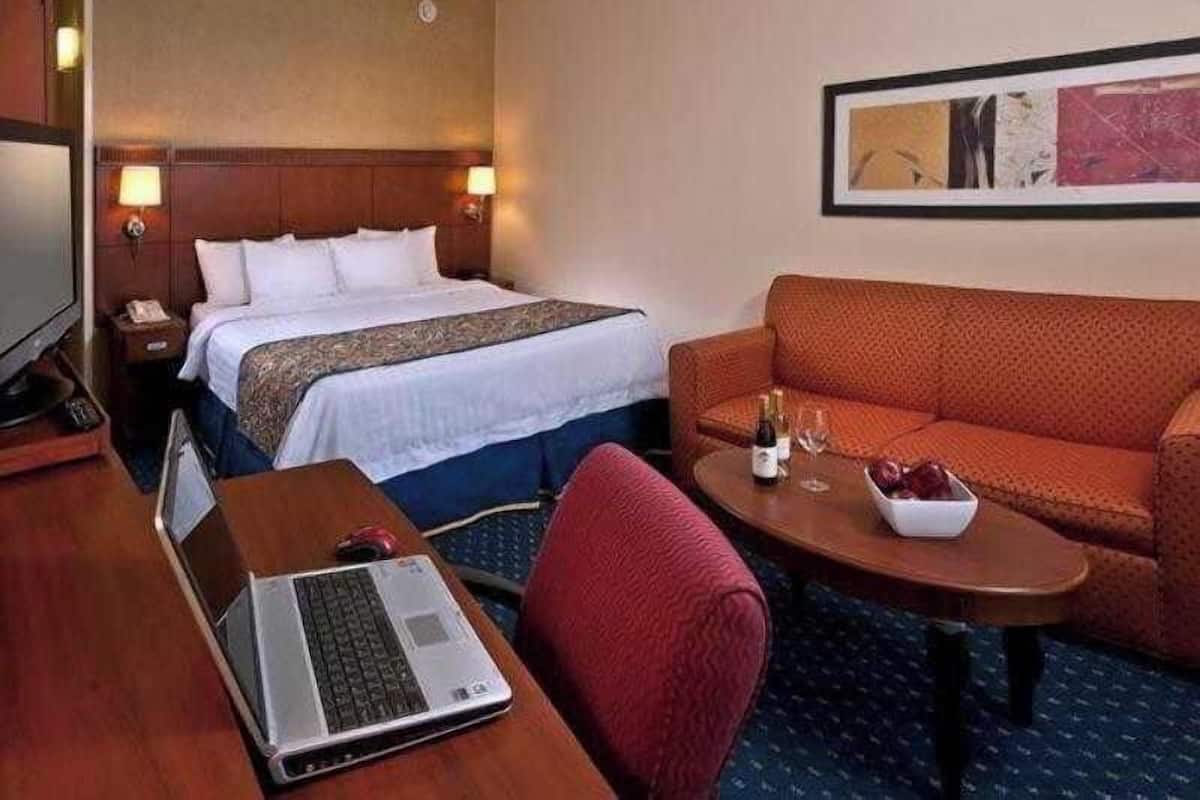 Ein Hotelzimmer mit einem Bett, einem Schreibtisch mit Laptop, einer Couch und einem kleinen Tisch mit Flasche und Gläsern.