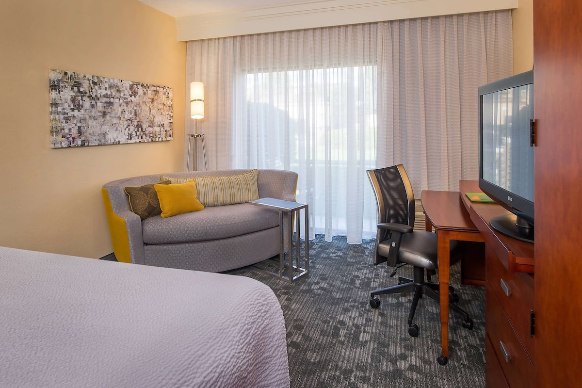 Ein Hotelzimmer mit einem Bett, einem Sofa mit gelben Kissen, einem Schreibtisch mit Computer, einem Fernseher und einem großen Fenster mit Vorhängen.