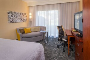 Una habitación de hotel con cama, sofá con cojines amarillos, escritorio con computadora, televisor y un ventanal con cortinas.