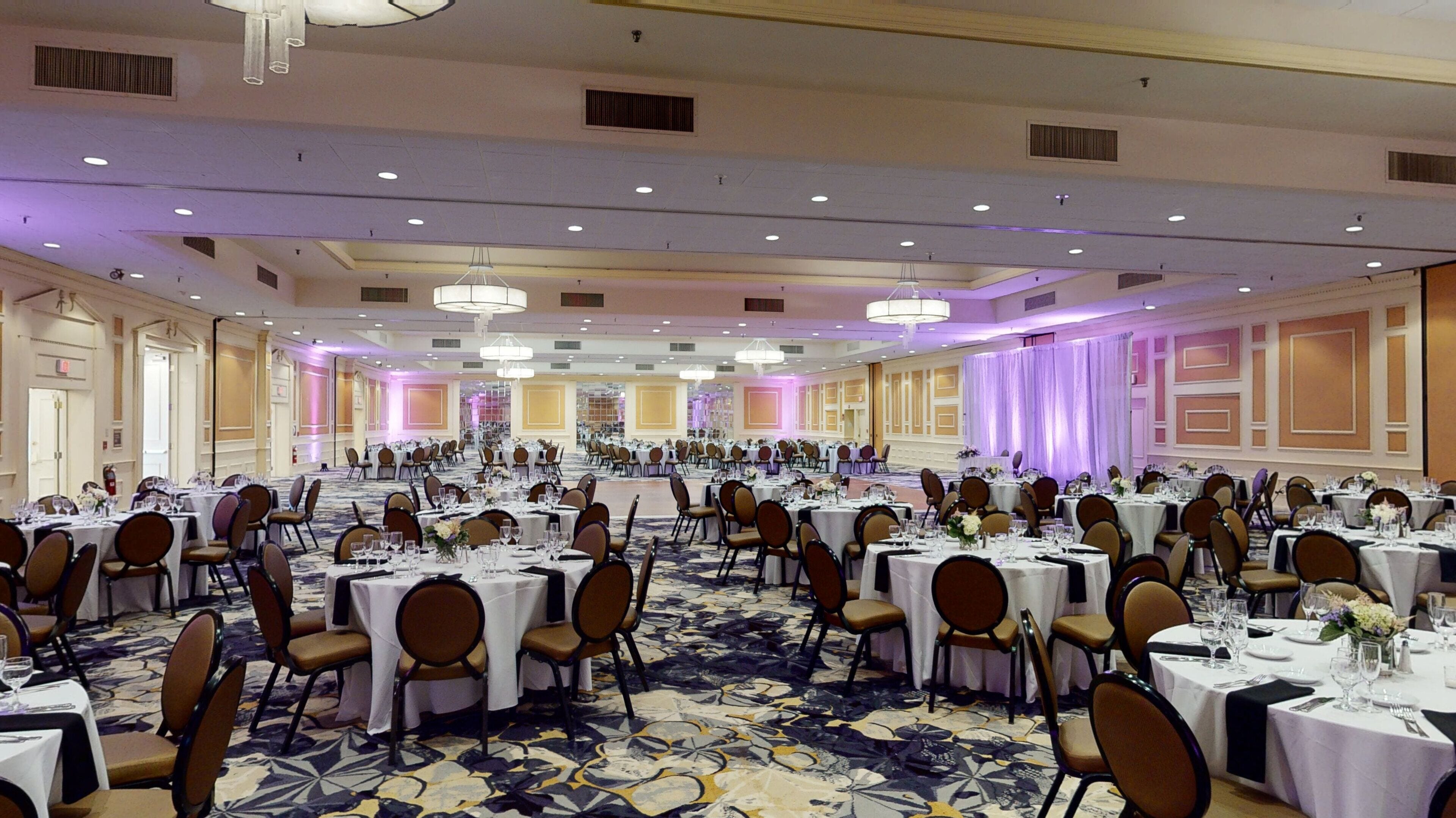 Une grande salle de banquet avec des tables rondes et des chaises disposées en rangées, un tapis à motifs et un éclairage encastré.