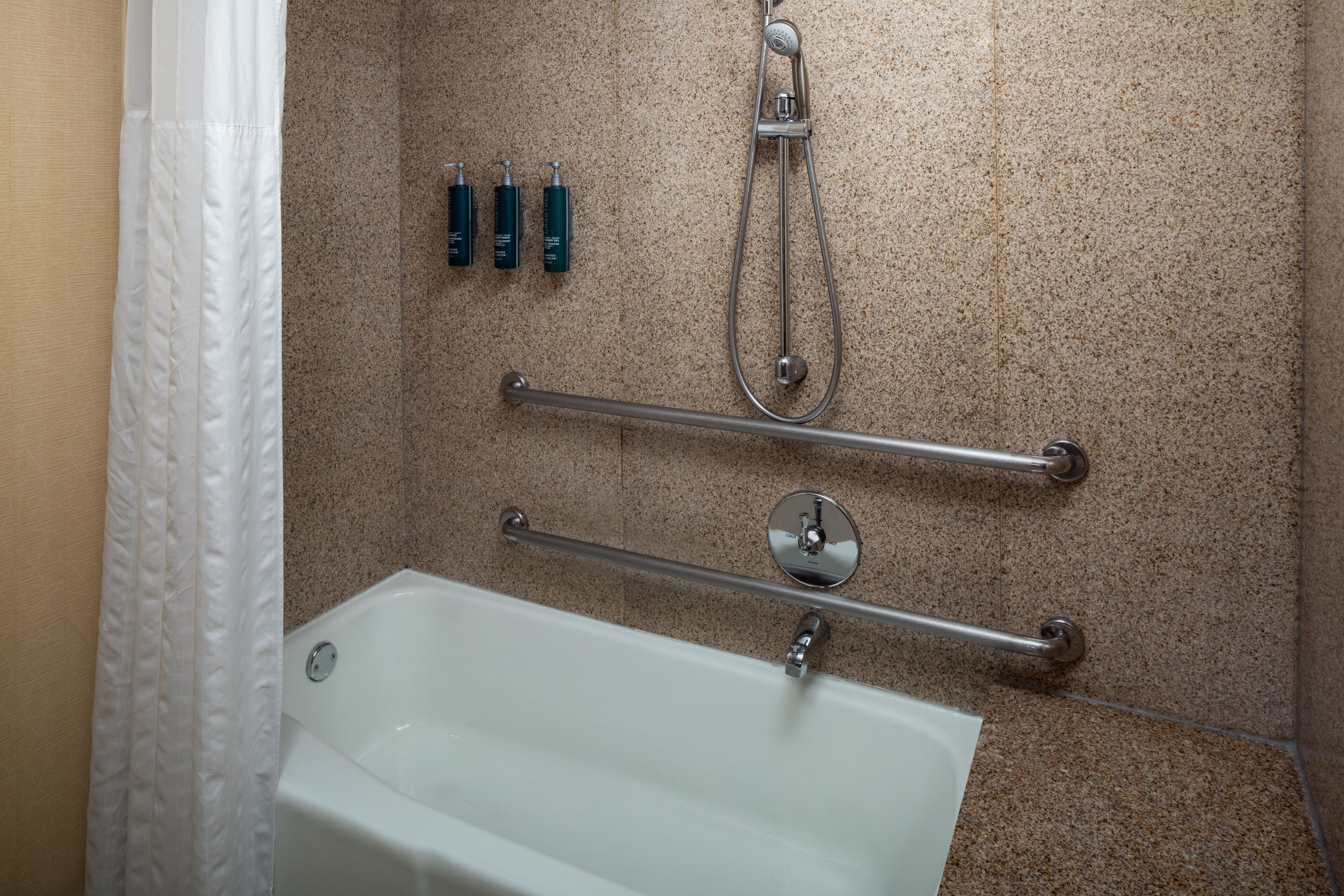 A bathroom with a white bathtub, grab bars, a showerhead, and three bottles of shampoo.