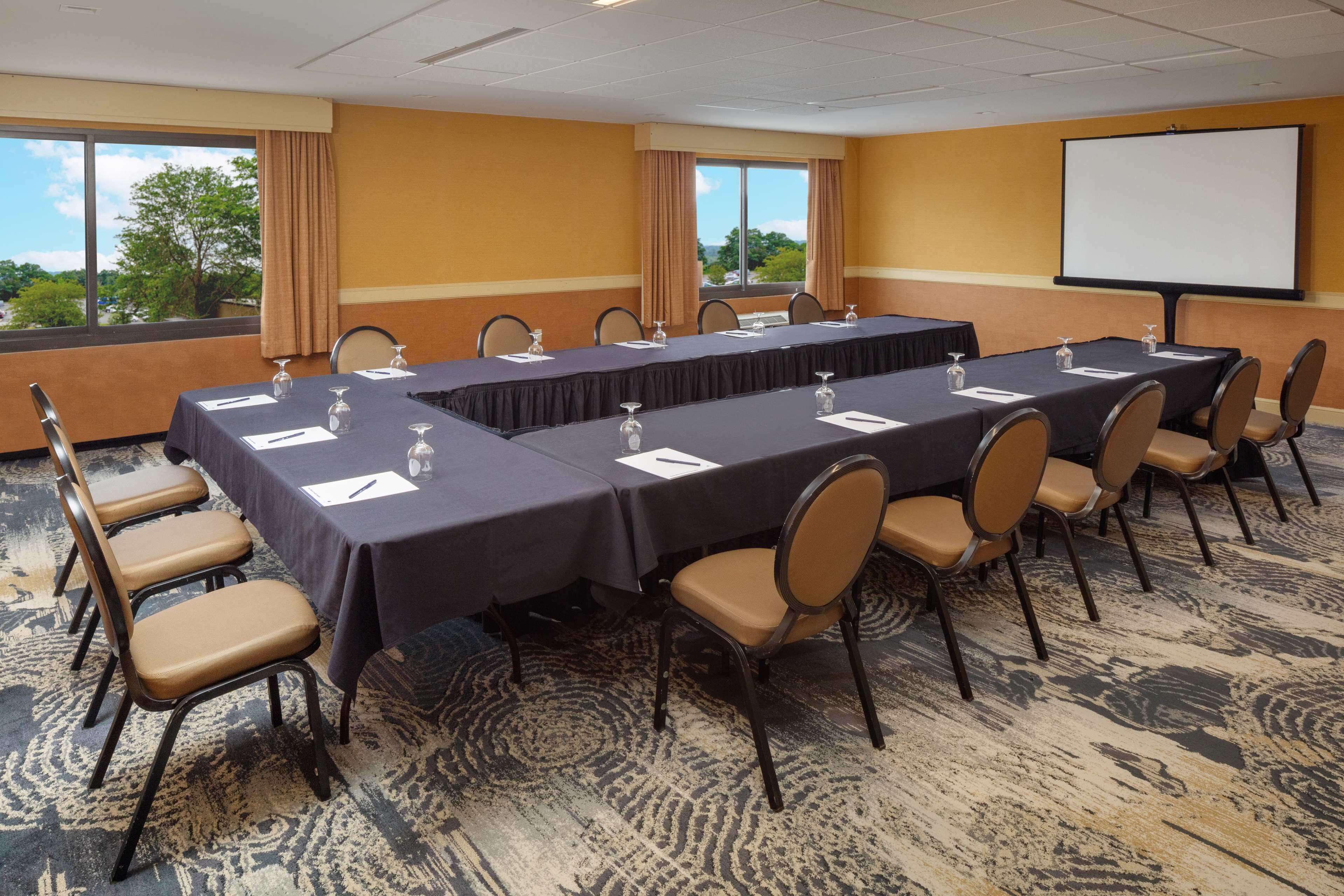 A conference room with a long table, chairs, a projector screen, and a view of trees through the windows.