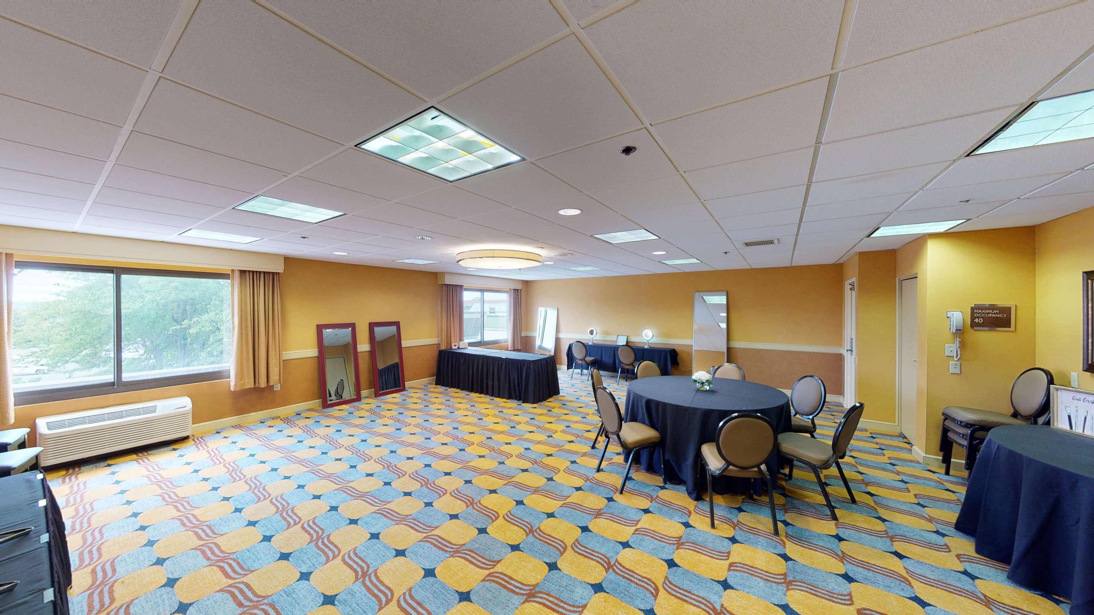 A conference room with a patterned carpet, multiple tables with chairs, large windows, and a ceiling with recessed lighting.