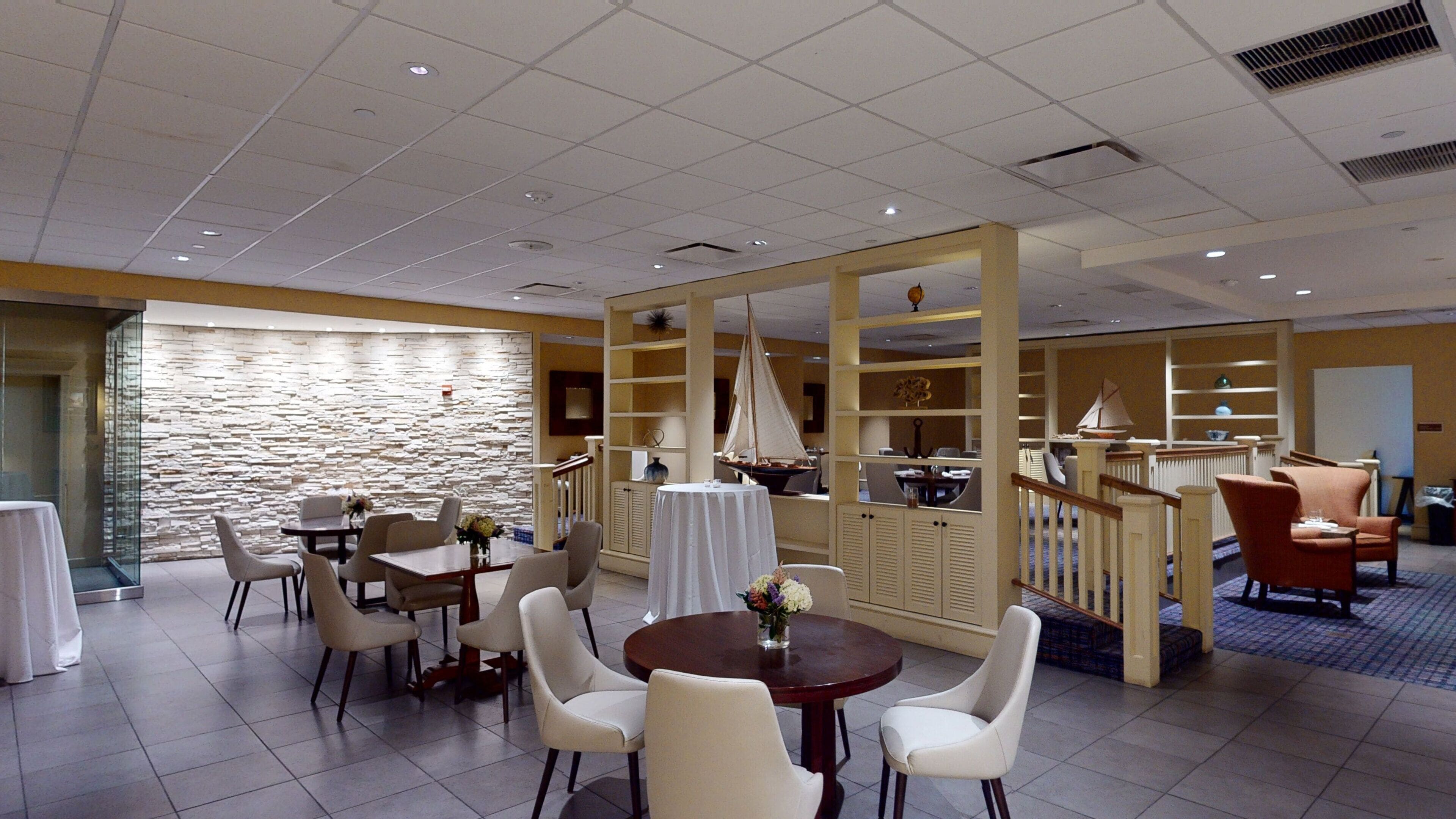 A hotel dining area with multiple tables and chairs, a stone wall, and a staircase leading to another level.