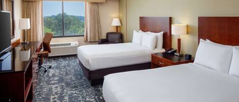 A hotel room with two beds, a desk, a chair, a TV, and a window with a view of the outdoors.