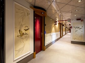 A hotel corridor with framed artwork on the walls, a red door, and a long hallway with a patterned carpet.