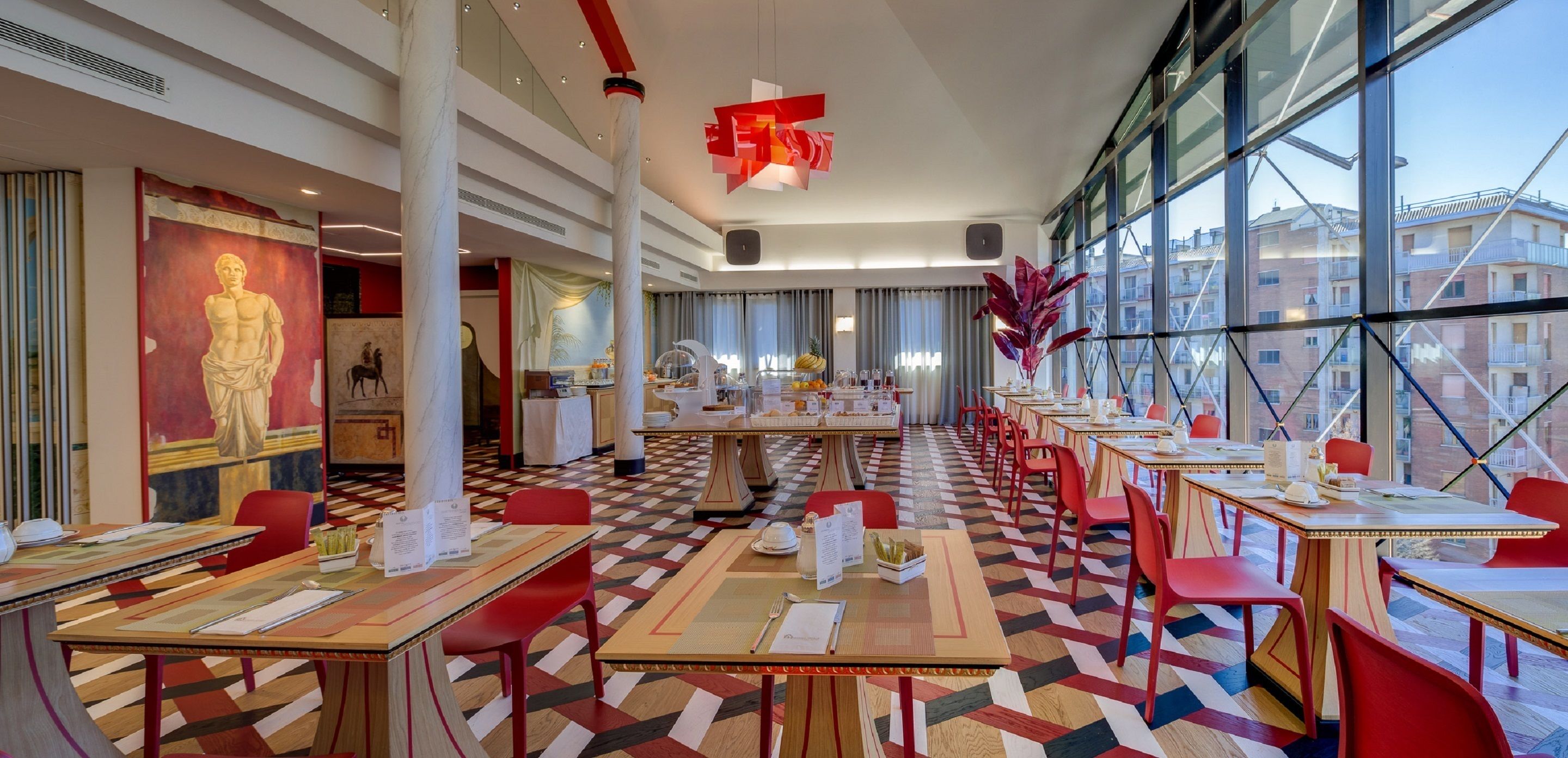 Une salle à manger d’hôtel avec de nombreuses tables et chaises, un sol à motifs et de grandes fenêtres donnant sur des bâtiments.