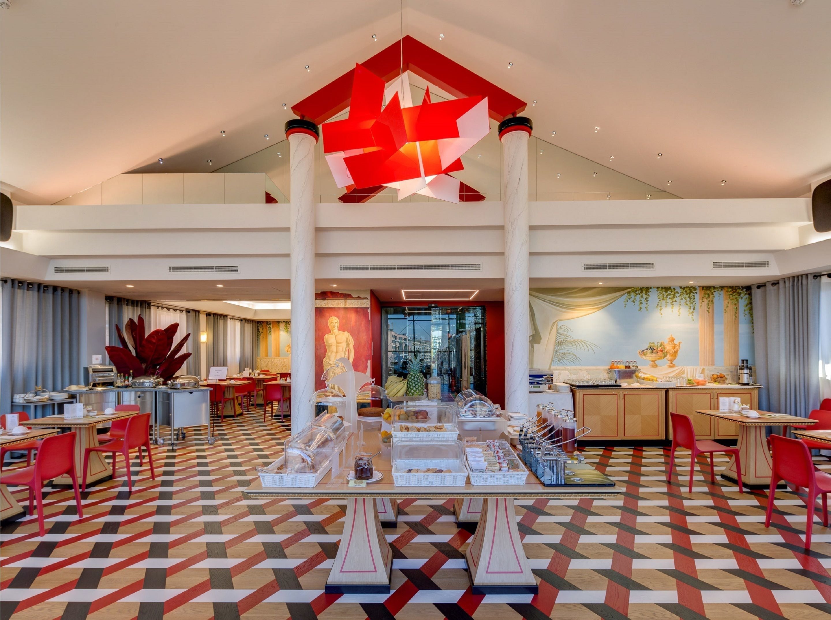 Un hall de hotel espacioso con diseño moderno, que cuenta con una zona de comedor central con una escultura geométrica en el techo, un suelo con diseño y vistas a la ciudad a través de amplias ventanas.
