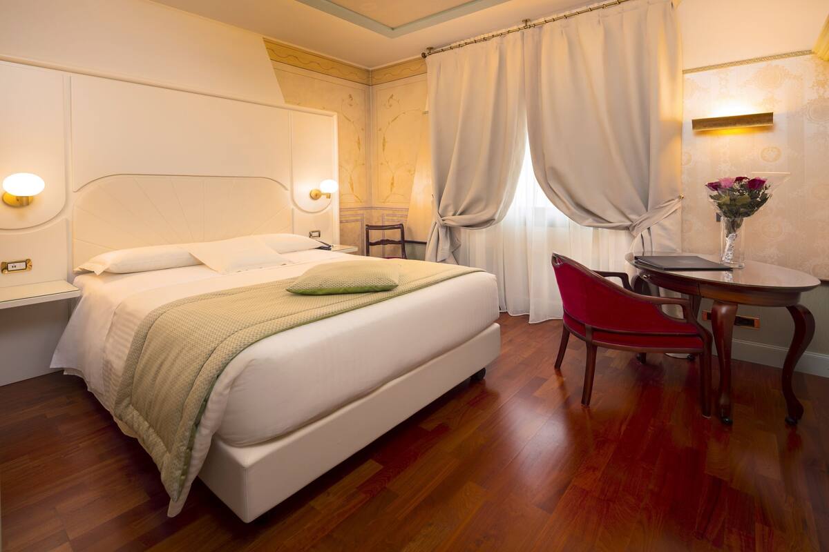 A hotel room with a bed, bedside table, chair, and a small table with a vase of flowers.