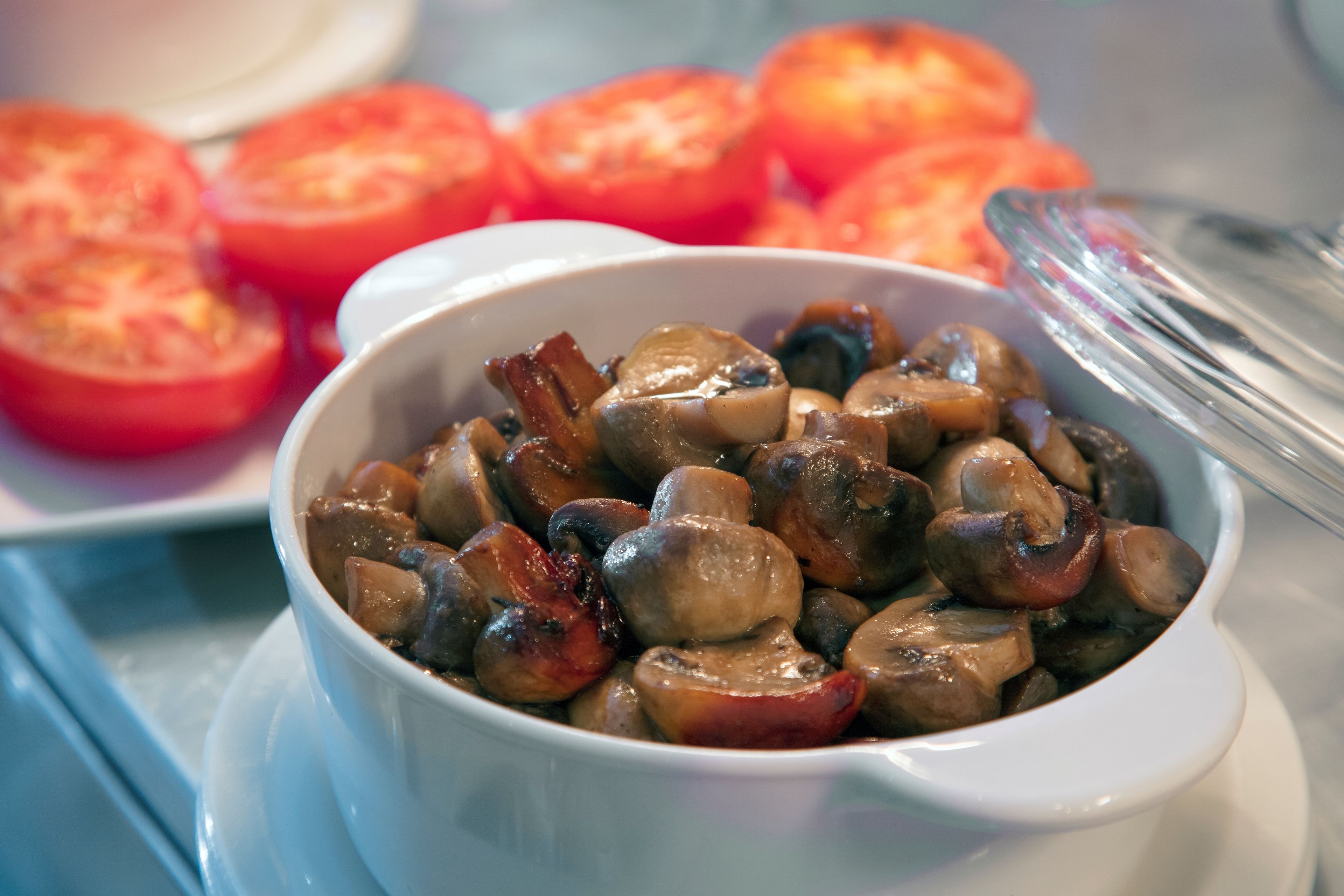A bowl of cooked mushrooms with a white bowl and a white plate with sliced tomatoes in the background.