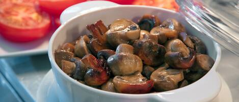 A bowl of cooked mushrooms with a white bowl and a white plate with sliced tomatoes in the background.