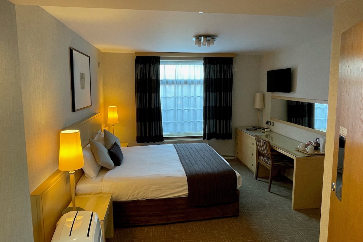 Une chambre d’hôtel équipée d’un lit, de lampes de chevet, d’un bureau, d’une chaise, d’une télévision et d’une fenêtre avec des rideaux.