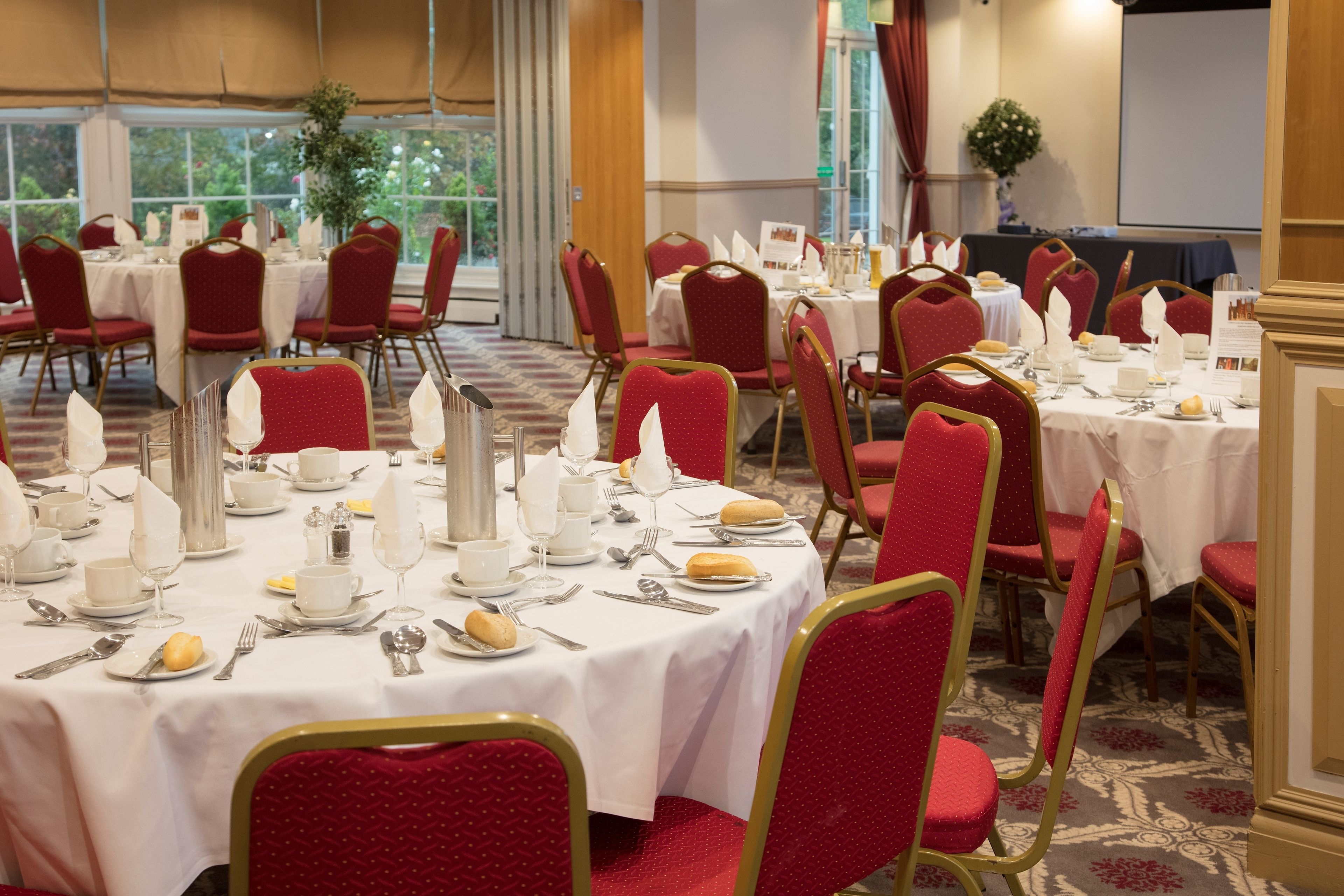 A banquet hall with round tables set for dining, red upholstered chairs, and a large window providing natural light.