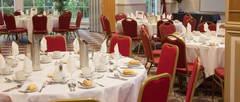 A banquet hall with round tables set for dining, red upholstered chairs, and a large window providing natural light.