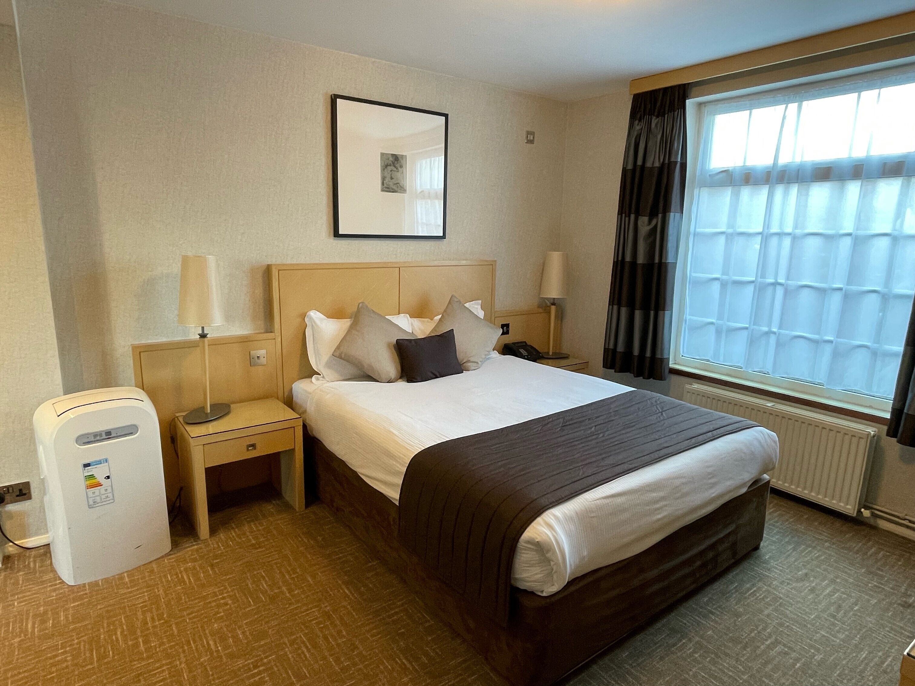 A hotel room with a bed, bedside tables, a heater, and a window with curtains.