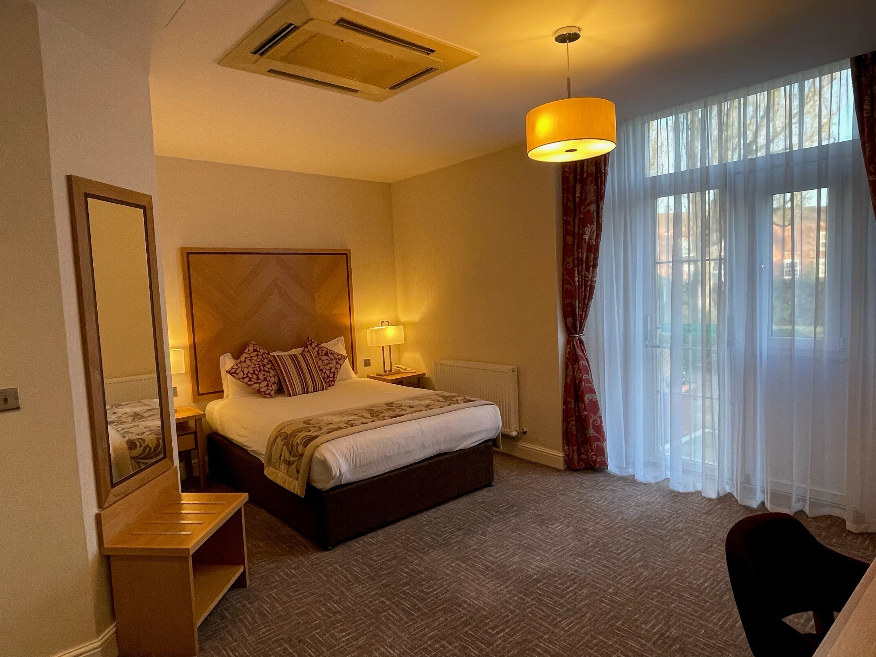 A hotel room with a bed, bedside table, lamp, and a view of the outdoors through large windows.
