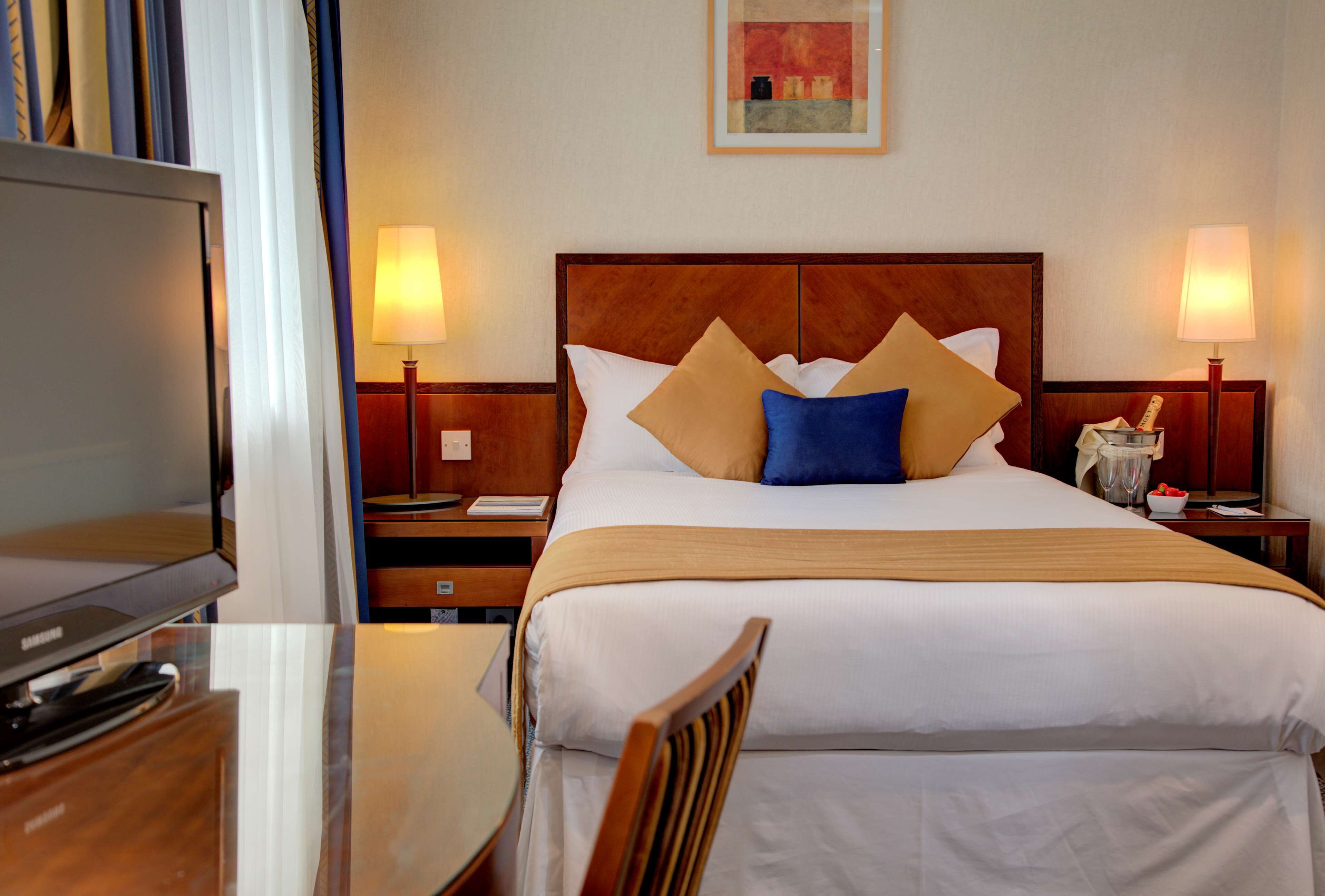 A hotel room with a bed, two bedside lamps, a television, a wooden chair, and a small table.