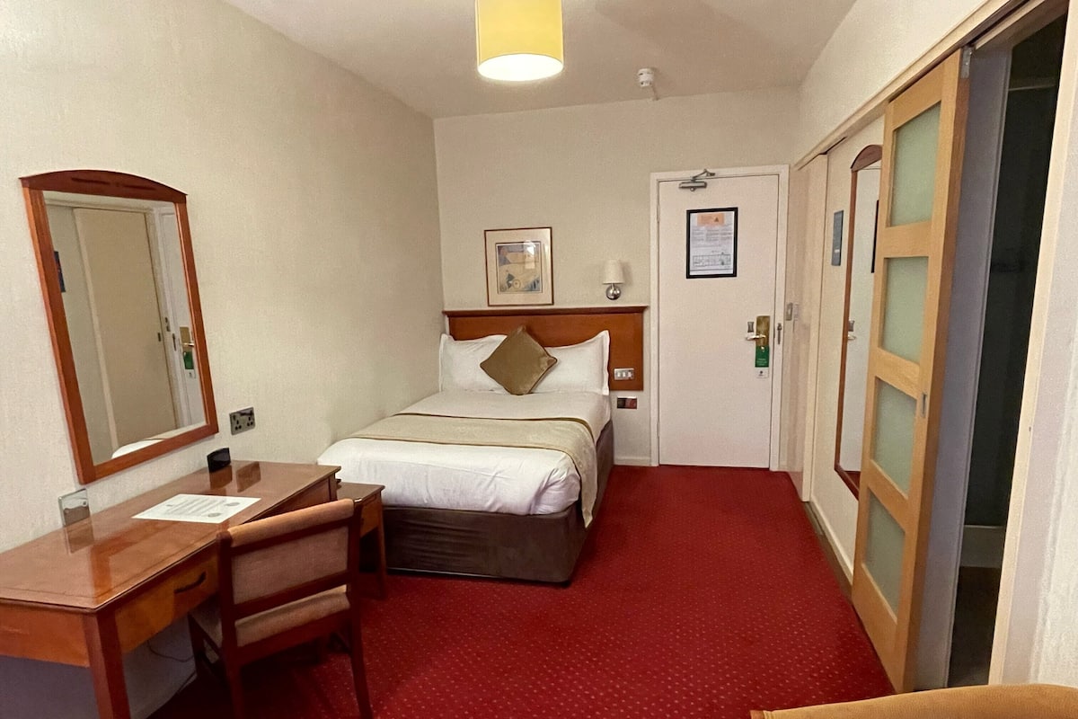 Une chambre d’hôtel avec un lit, un bureau, une chaise, un miroir et une porte.