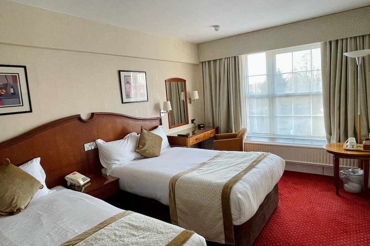 Une chambre d’hôtel avec deux lits, un bureau, une chaise, une fenêtre avec des rideaux et une lampe.