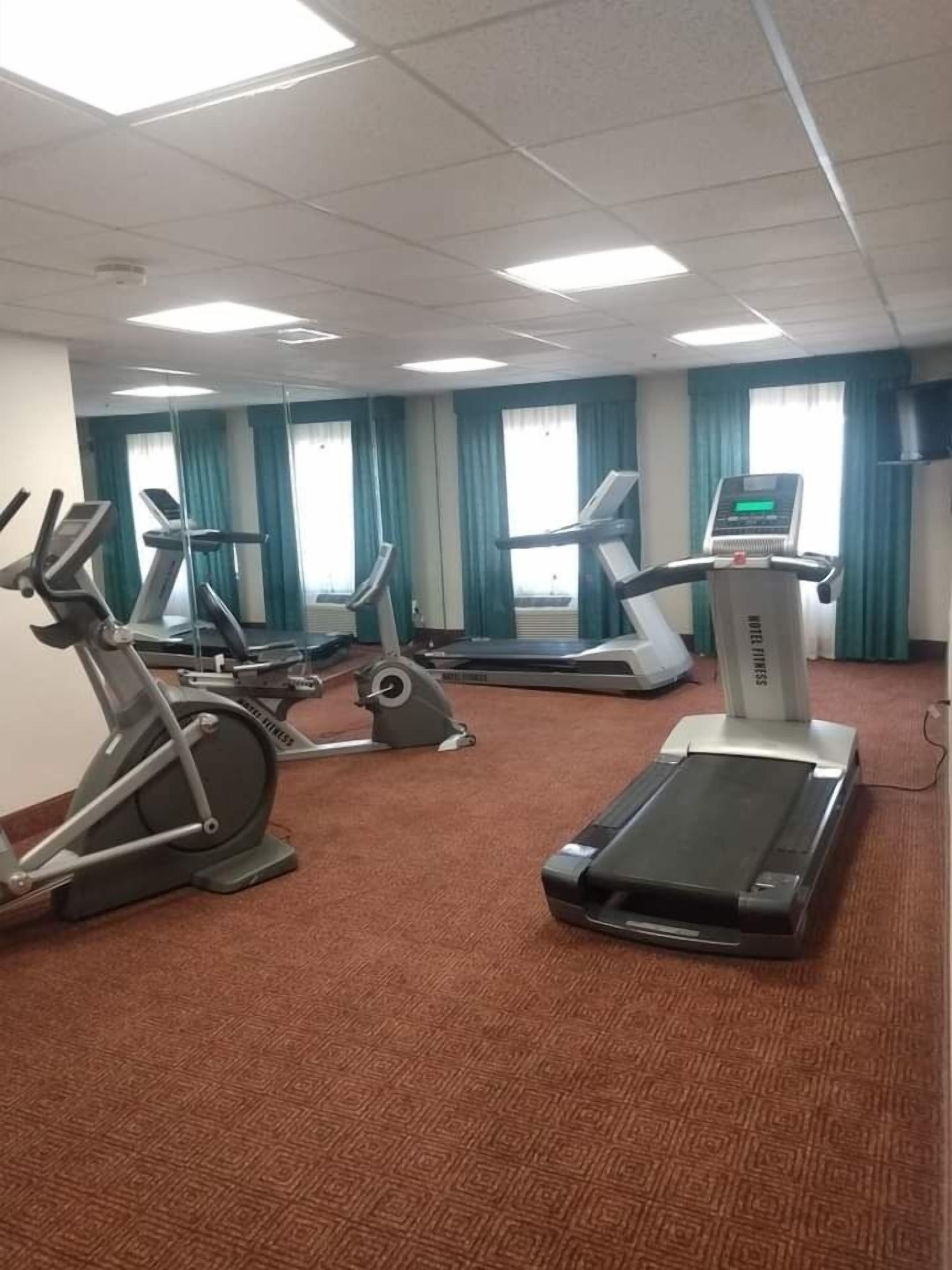 A gym with multiple treadmills and stationary bikes, carpeted floor, and large windows with curtains.