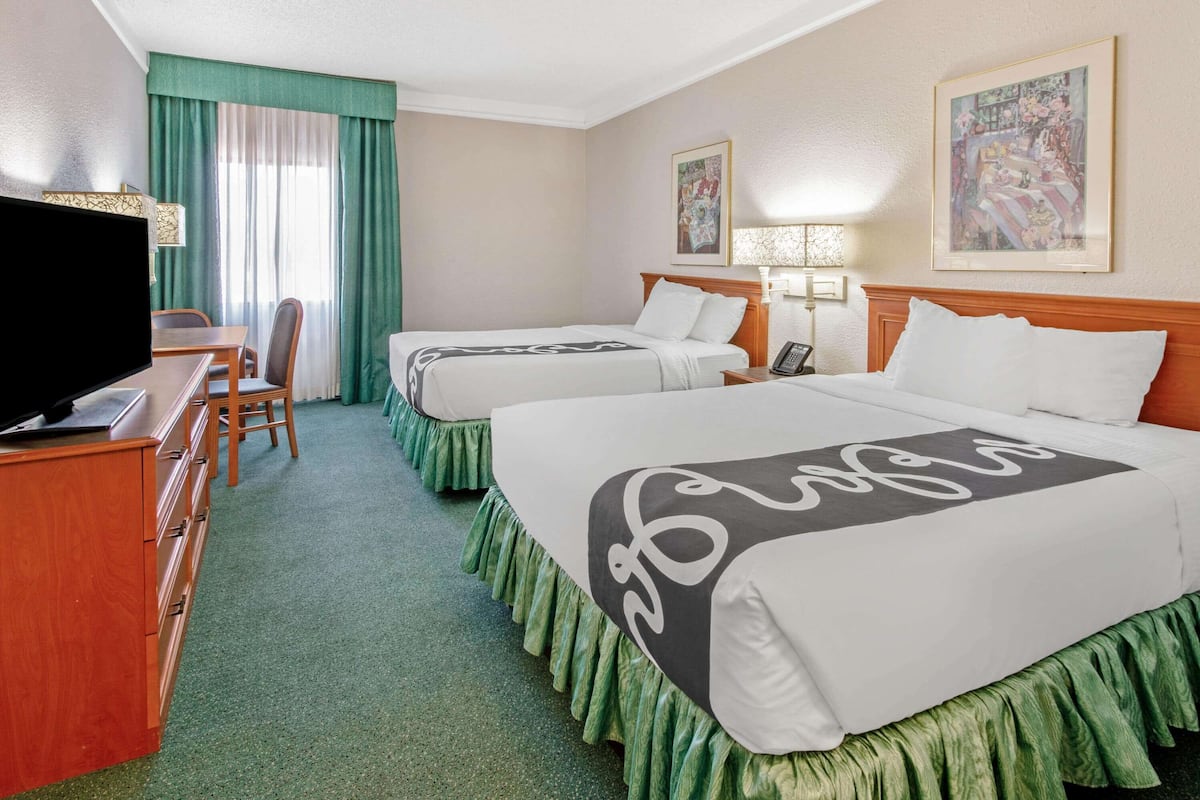 Ein Hotelzimmer mit zwei Betten, einem Schreibtisch mit Fernseher, einem Sessel und einem Fenster mit Vorhängen.