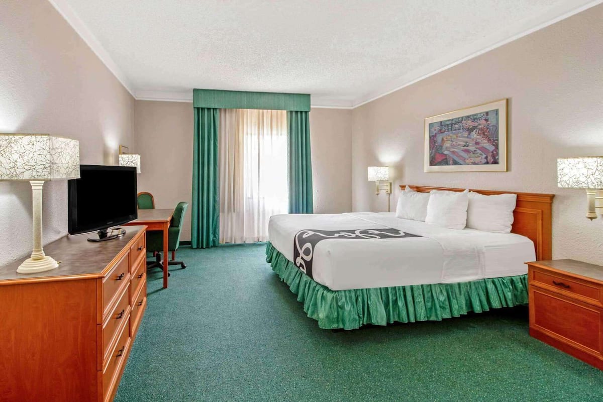 Ein Hotelzimmer mit einem Bett, einem Schreibtisch mit Fernseher, einem Sessel, einem Schminktisch und einem Bild an der Wand.