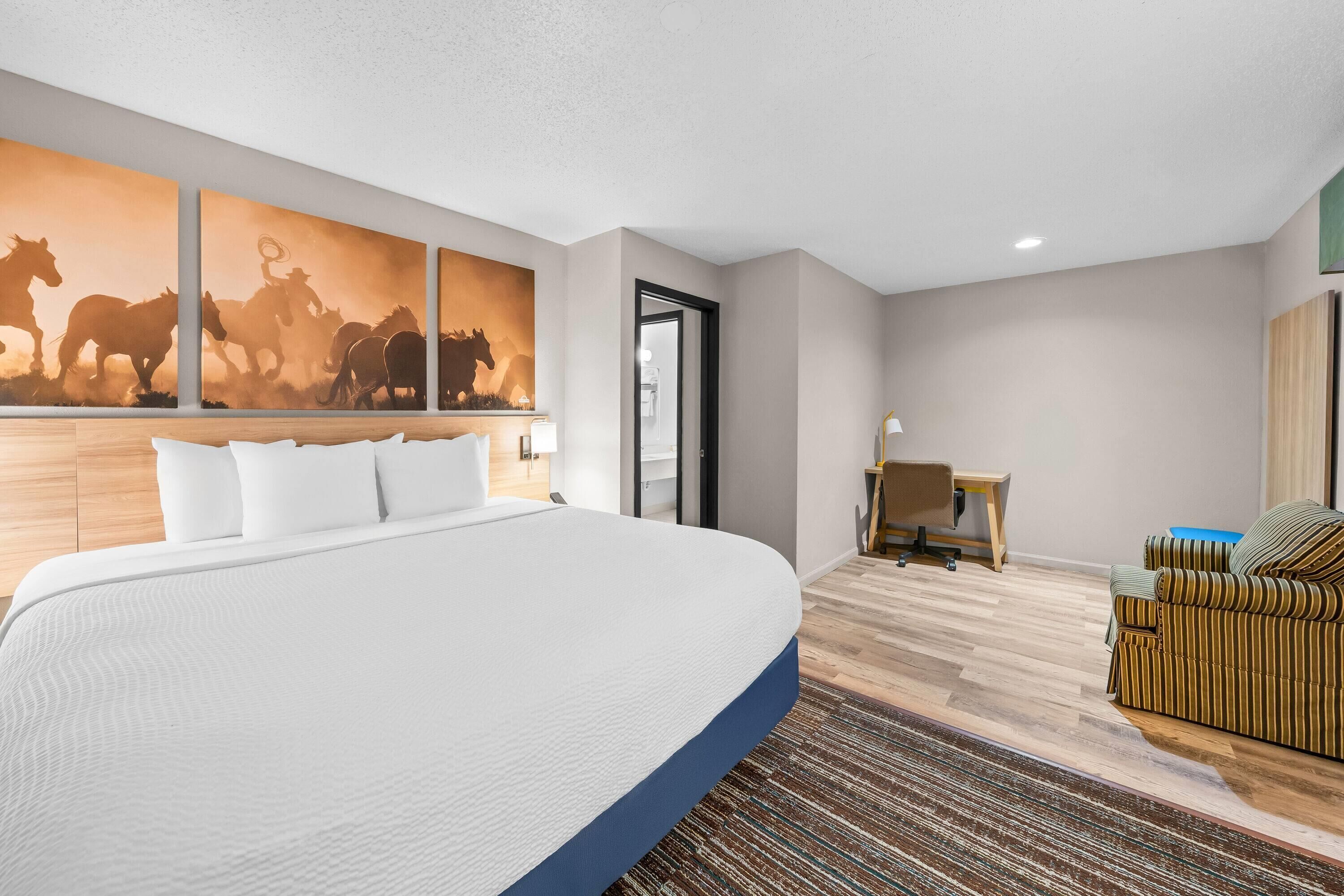 A hotel room with a large bed, a desk, a chair, and a wall-mounted painting of horses.