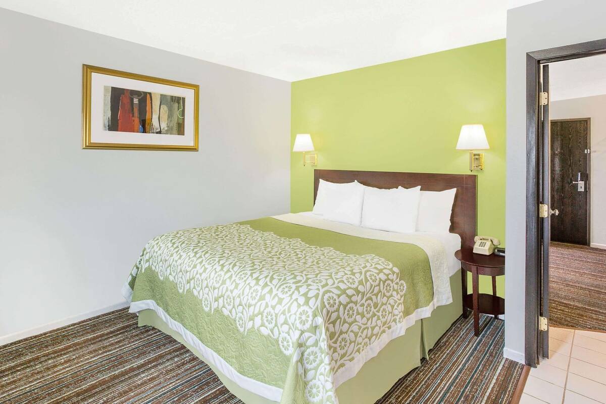 A hotel room with a bed, two bedside lamps, a framed picture on the wall, and a door leading to another room.