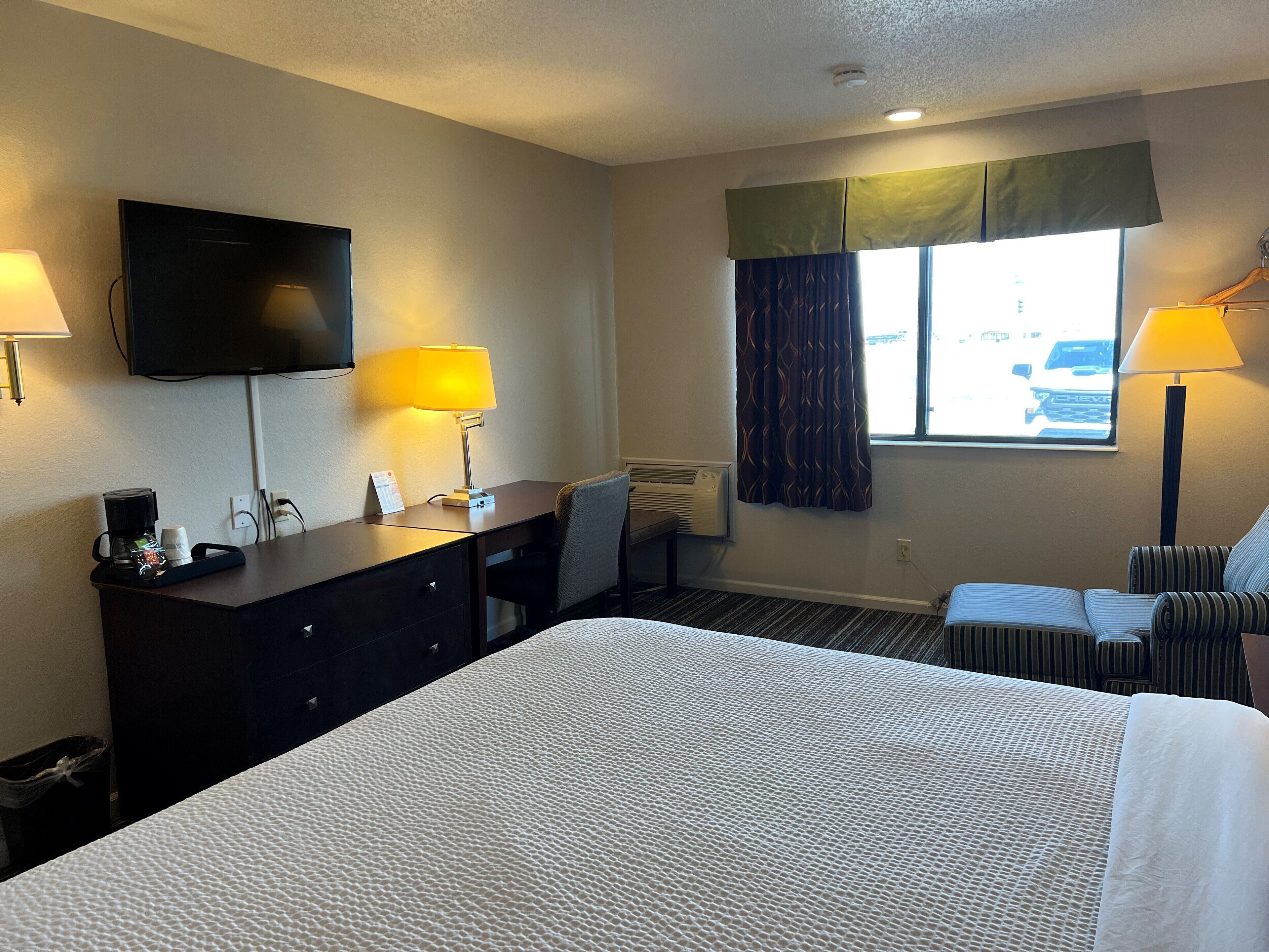 A hotel room with a bed, desk, chair, lamps, TV, and a window with curtains.