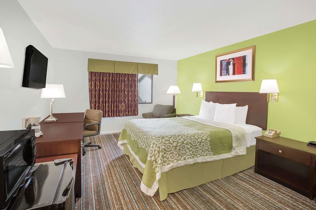 A hotel room with a bed, two bedside lamps, a desk with a lamp, a chair, a television, and a painting on the wall.