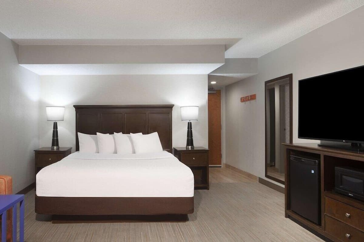 A hotel room with a large bed, two bedside tables with lamps, a flat-screen TV, a small refrigerator, and a wooden cabinet.