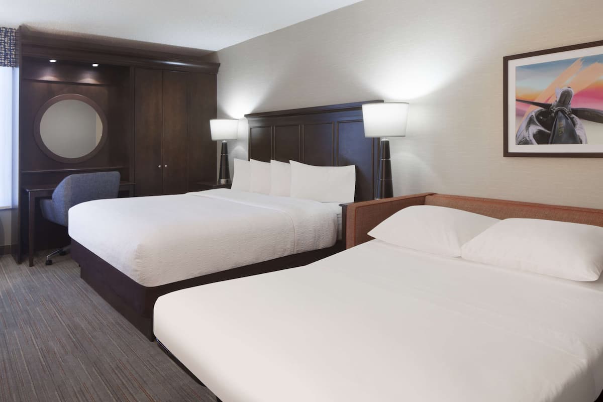 A hotel room with two beds, a desk, a chair, a round mirror, and a painting on the wall.