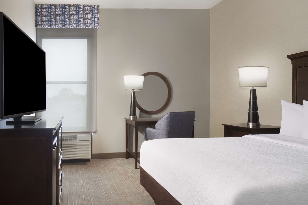 A hotel room with a bed, a TV, two bedside lamps, a desk with a chair, and a window with a view.