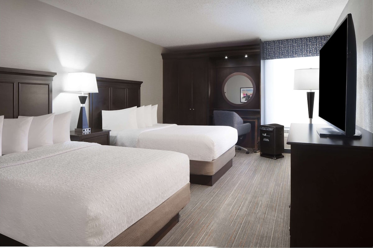 A hotel room with two beds, a desk, a chair, a television, and a large window.