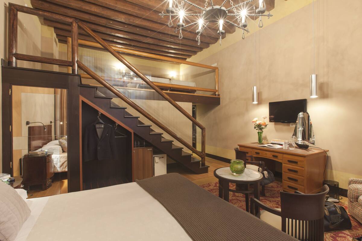 Habitación de hotel con cama, escritorio, silla, mesita, espejo, cómoda, lámpara, reloj, televisor y una escalera que lleva a un nivel superior.