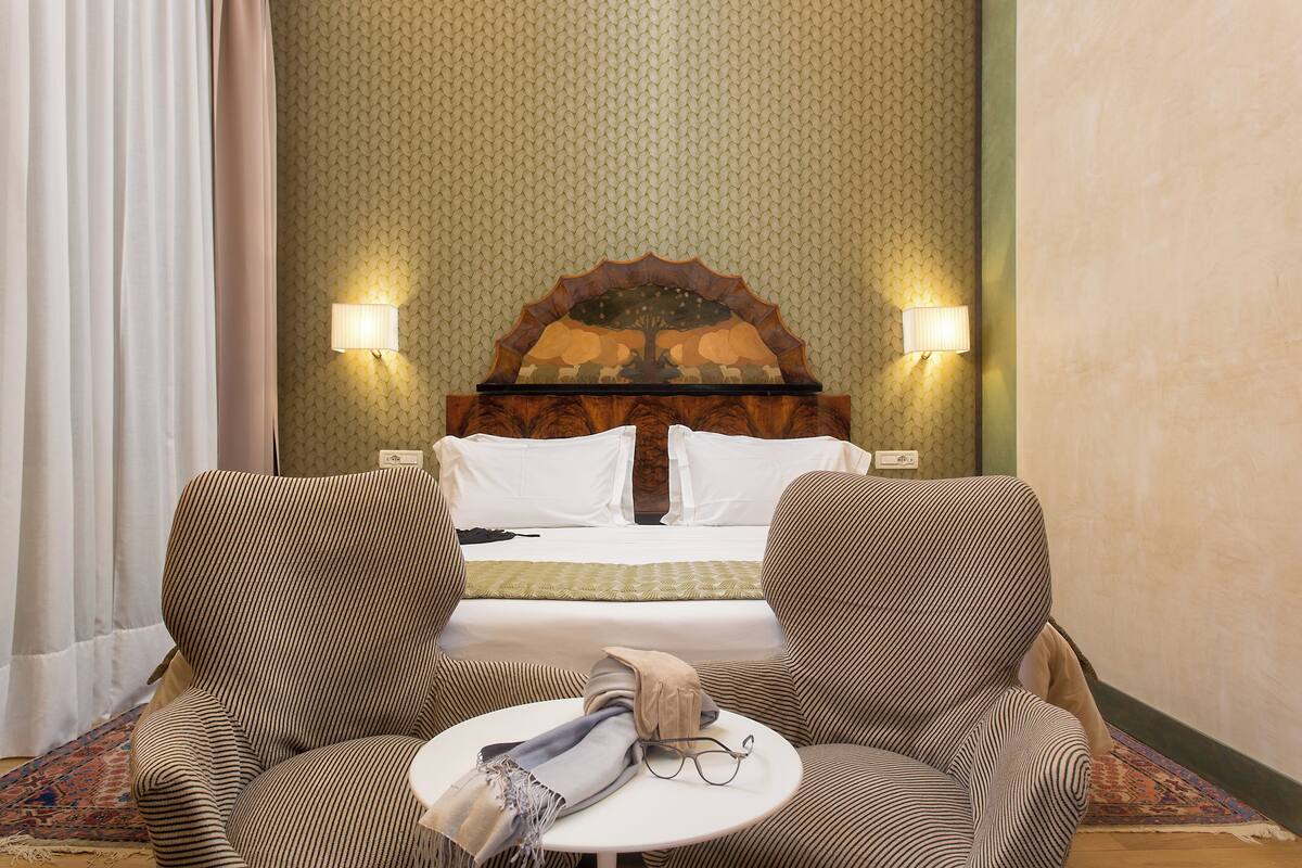 A hotel room with a bed, two armchairs, a small table, and a decorative wall panel.