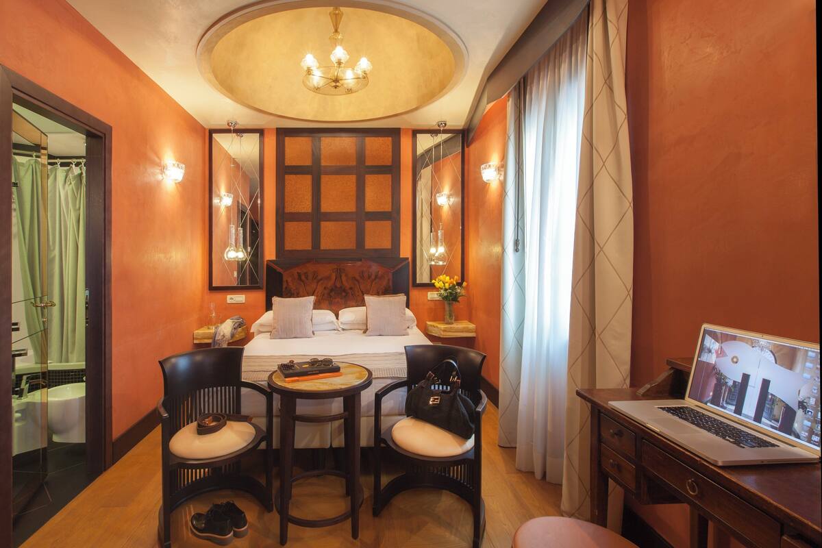 A hotel room with a bed, two chairs, a desk with a laptop, a mirror, and a chandelier.