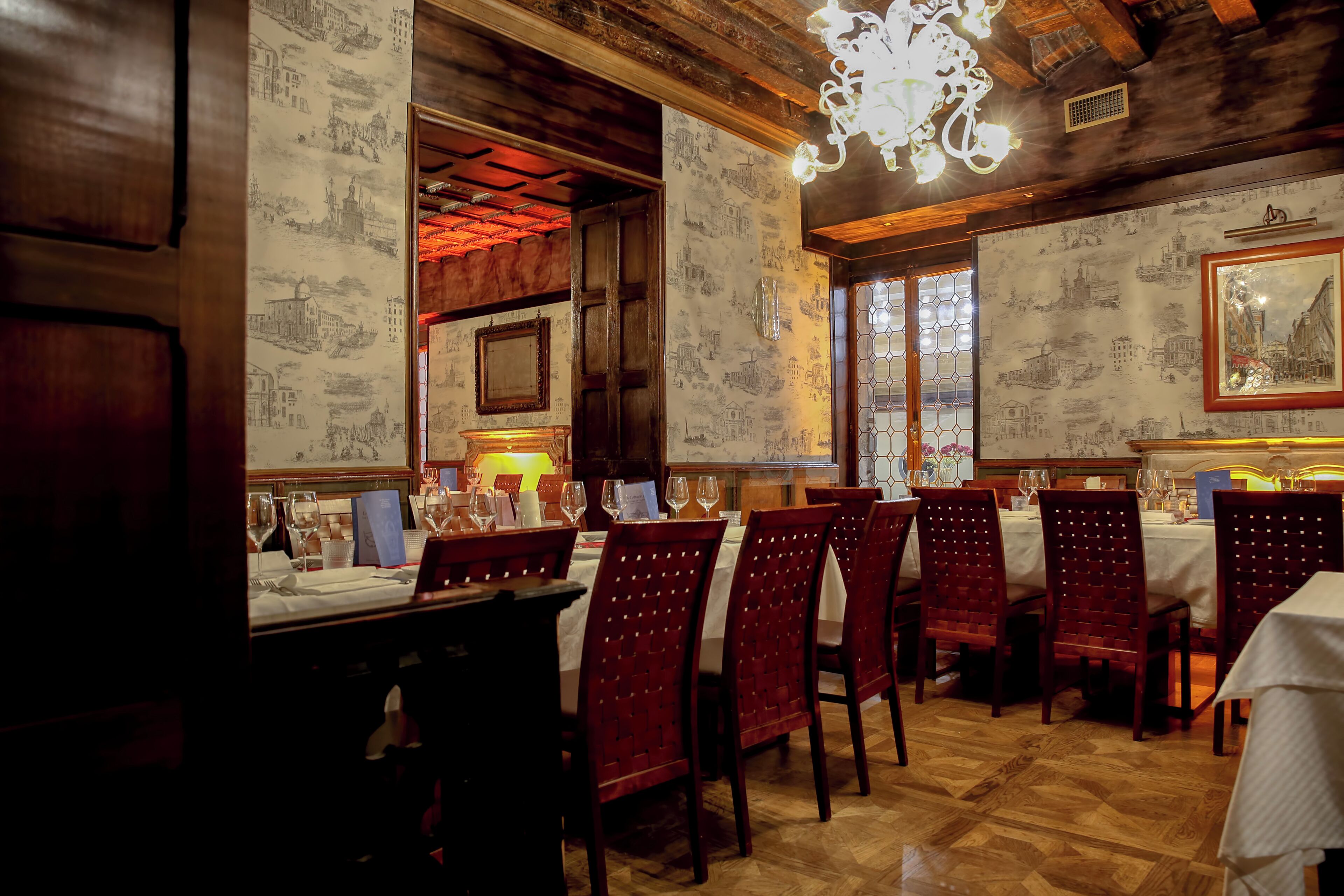A restaurant with wooden interiors, patterned wallpaper, and a chandelier. Tables are set with wine glasses and menus.