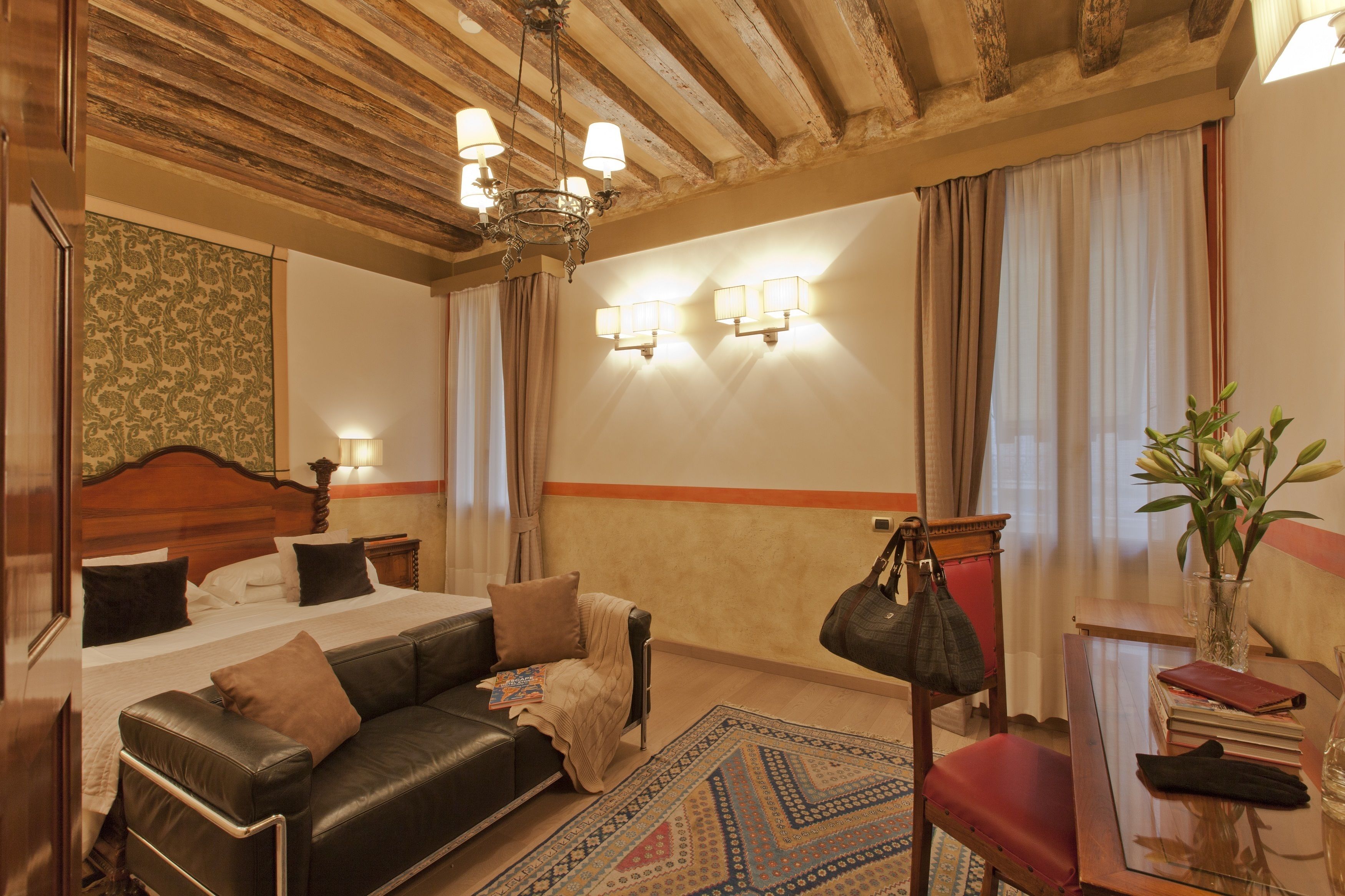 A hotel room with a bed, a leather sofa, a wooden headboard, a chandelier, wall sconces, a red chair, a small table with a vase of flowers, and a patterned rug.