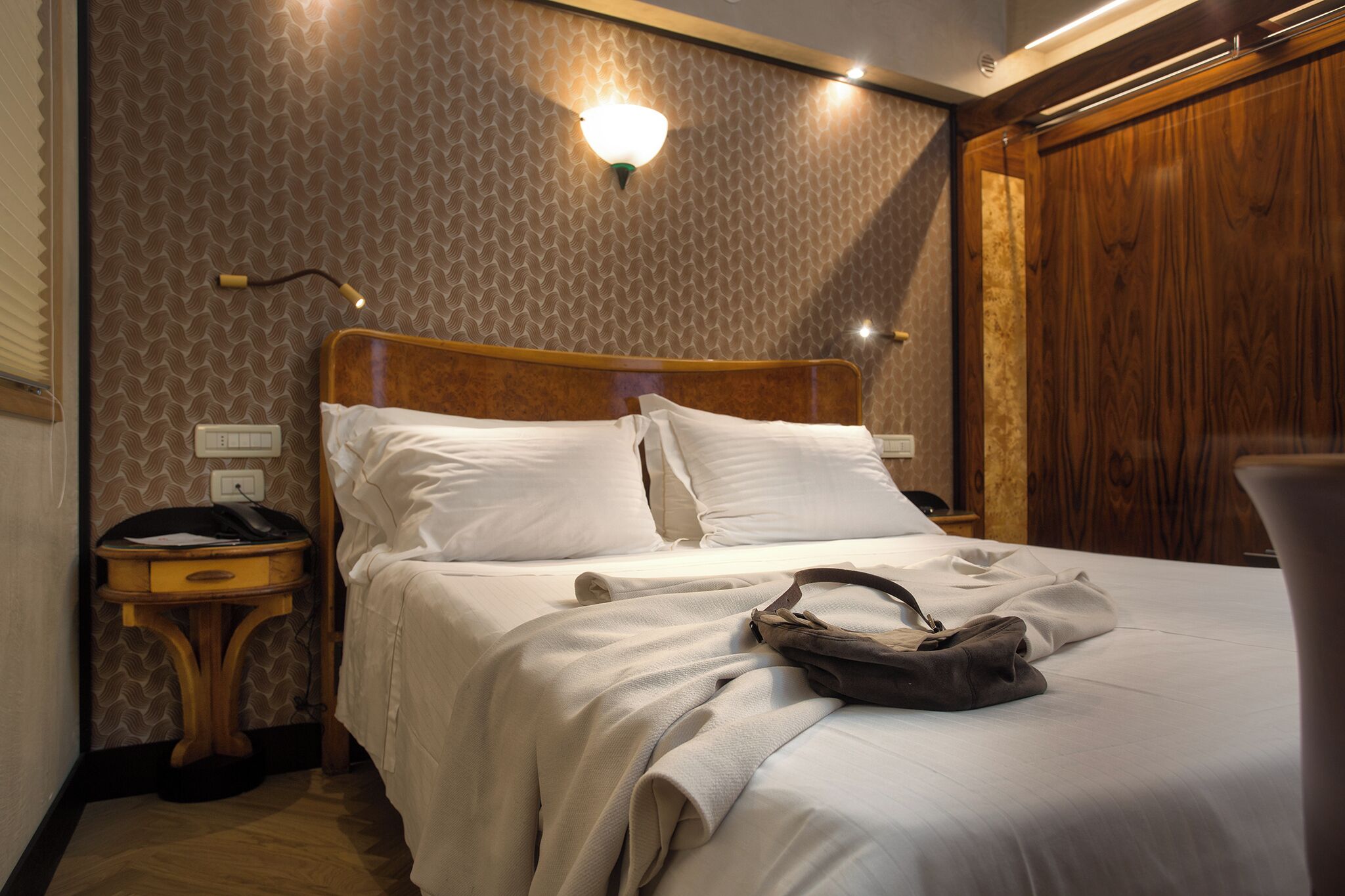 A neatly made bed with white linens, a wooden headboard, a bedside table with a lamp, and a wall with a patterned wallpaper.