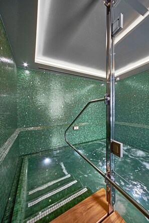 A modern bathroom with green mosaic tiles, a glass shower enclosure, and a wooden bench.