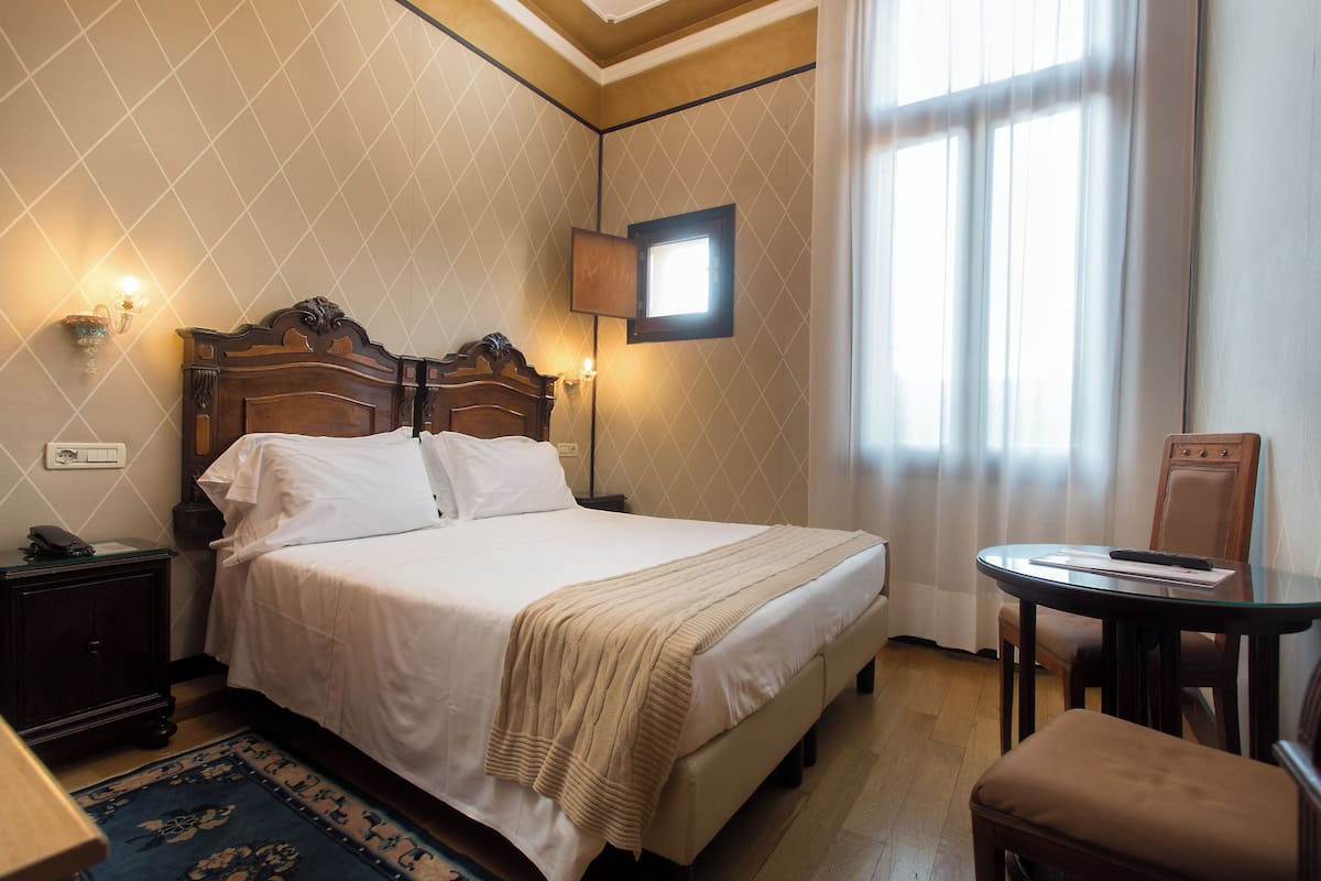 Una cama bien hecha con cabecera, una mesita de noche con lámpara, una pequeña mesa redonda con una silla y una ventana con cortinas.